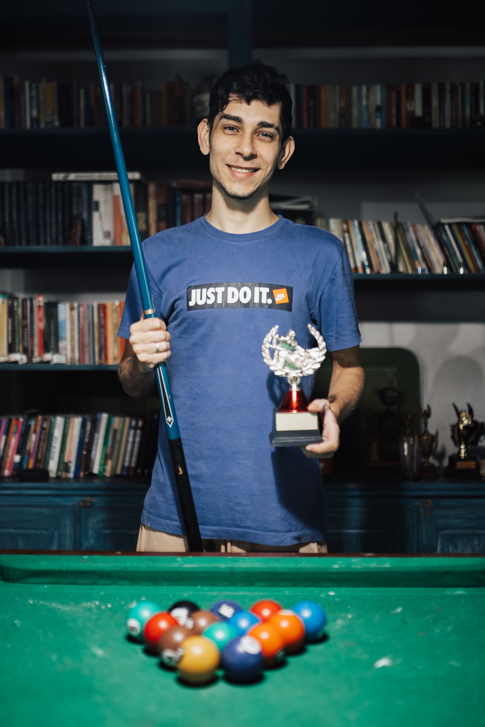 Man holding a pool cue and a trophy beside a pool table.